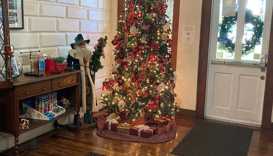 A festively decorated Christmas tree with red accents stands next to a life-size figure dressed as a Christmas character in an indoor setting.