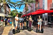 A group of four people are smiling and standing on Segways on a pedestrian street with outdoor cafes and palm trees in the background.