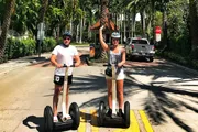 Two people wearing helmets are standing on Segways on a sunny, palm-lined road, with the woman raising her arm high as if gesturing or signaling.