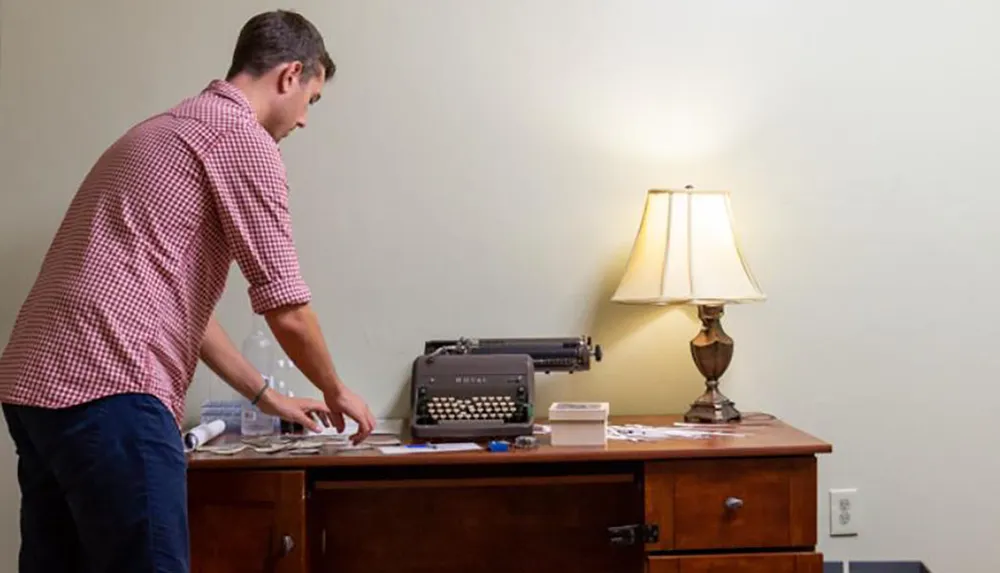 A man is standing at a wooden desk with a vintage typewriter on it alongside a lamp and some papers in a room with plain walls