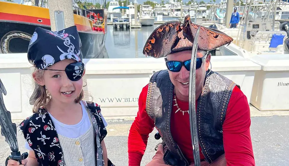 A child and an adult dressed in pirate costumes are smiling with a marina in the background