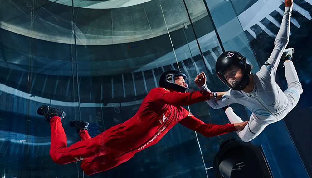 Two people are engaging in indoor skydiving wearing protective helmets and suits as they float in a vertical wind tunnel