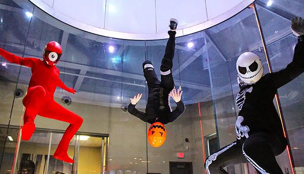 Three individuals in costume are simulating flight in a vertical wind tunnel with one dressed as Jack Skellington from The Nightmare Before Christmas another in a superhero-like outfit and the third person upside down holding a Halloween-themed pumpkin bucket
