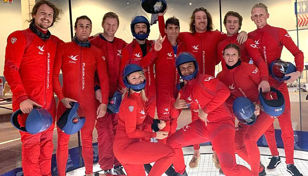 A group of people in matching red jumpsuits and blue helmets are posing for a photo likely before or after participating in an indoor skydiving or similar aerial activity