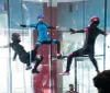 Three individuals in full-body suits and helmets are indoors skydiving in a vertical wind tunnel with an onlooker visible in the background