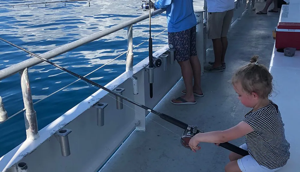 A young child is seated on the deck of a boat holding a fishing rod with other individuals standing nearby possibly engaged in a fishing activity