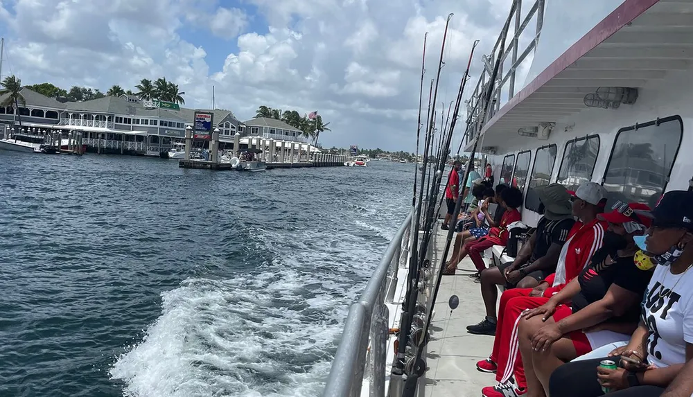 A group of people is seated along the railing of a fishing boat with rods lined up ready for a day of fishing with a waterfront building complex in the background