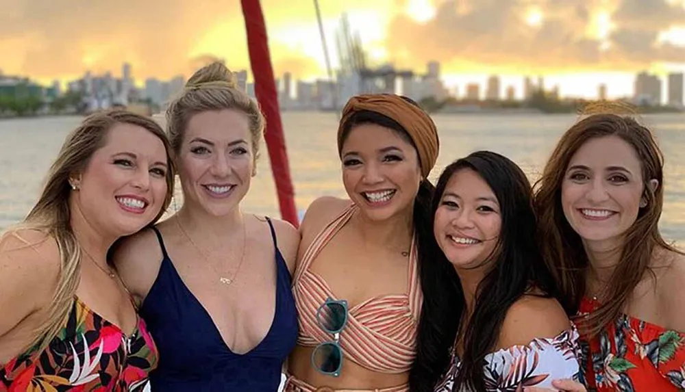 Five smiling women pose closely together for a photograph with a blurry sunset and city skyline in the background suggesting they are enjoying a social outing