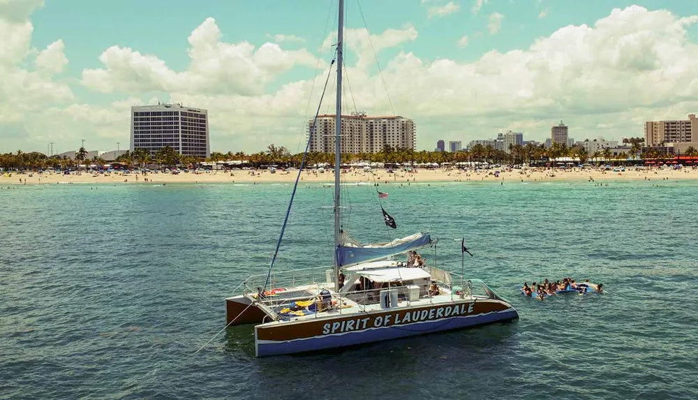 A catamaran named Spirit of Lauderdale is anchored near a bustling beach with a group of people swimming nearby in clear blue waters under a sunny sky adorned with fluffy clouds