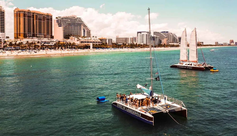 The image shows two catamarans with groups of people aboard sailing near a sunlit beachfront lined with palm trees and high-rise buildings
