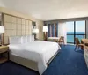 The image shows a well-appointed hotel room with a large bed modern furniture and a balcony overlooking a blue ocean