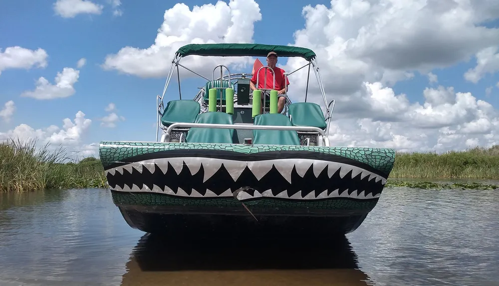 A person is standing on a pontoon boat designed to look like an alligator with its jaws open floating on calm water under a partly cloudy sky