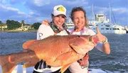 Two smiling women are holding a large fish on a boat with waterfront properties and yachts in the background.