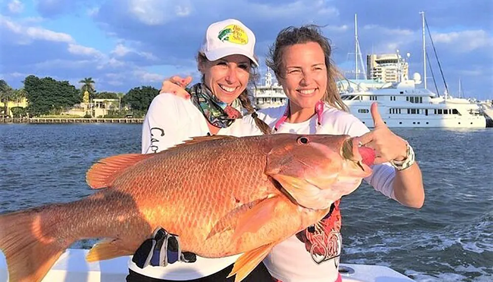 Two smiling women are holding a large fish on a boat with waterfront properties and yachts in the background