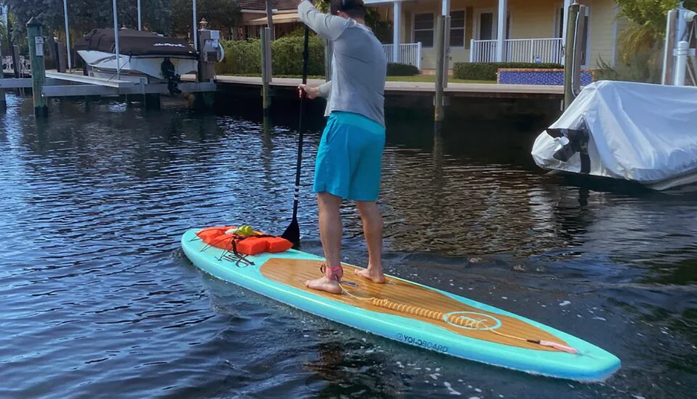 A person is stand-up paddleboarding on calm water near docked boats and waterfront houses