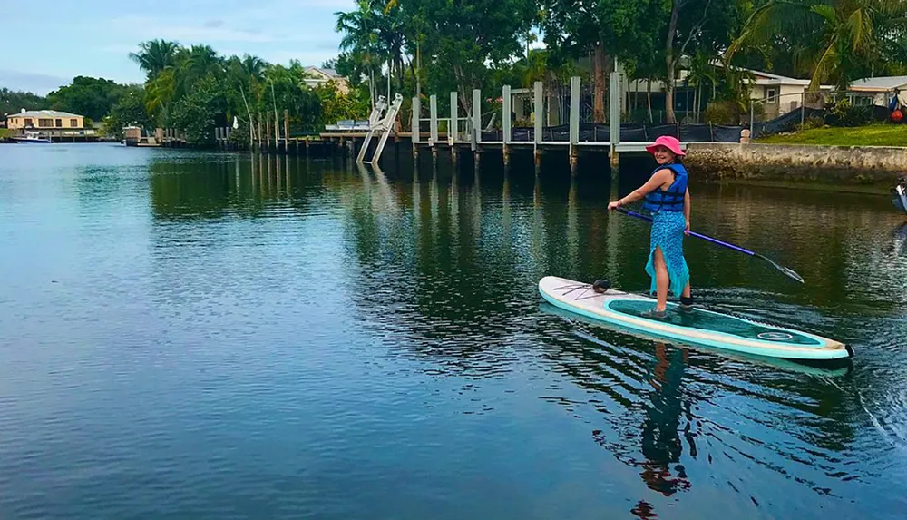 A person is stand-up paddleboarding on calm water with residential properties in the background