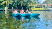 Three individuals are enjoying their time on a kayak in a scenic waterway surrounded by palm trees and waterfront properties.