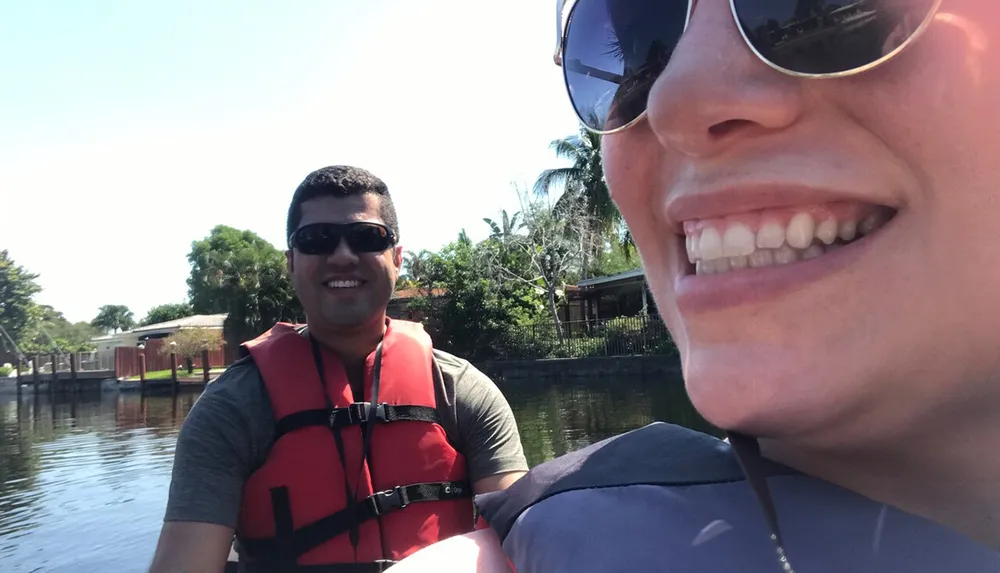 Two people are enjoying a sunny day on the water with one wearing a red life jacket and sunglasses and the other taking a selfie capturing part of their smiling face in the foreground