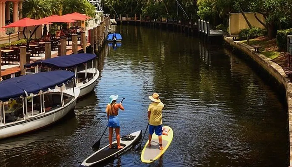 Two people are paddleboarding on a peaceful waterway flanked by boats and a dining area with red umbrellas