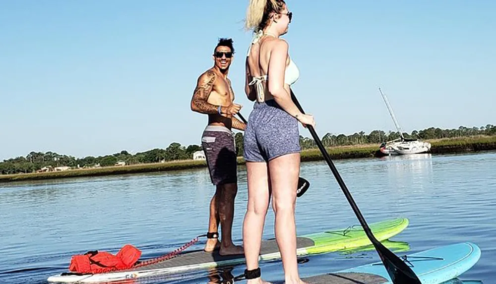 Two people are standing on paddleboards on a calm body of water with a boat visible in the background