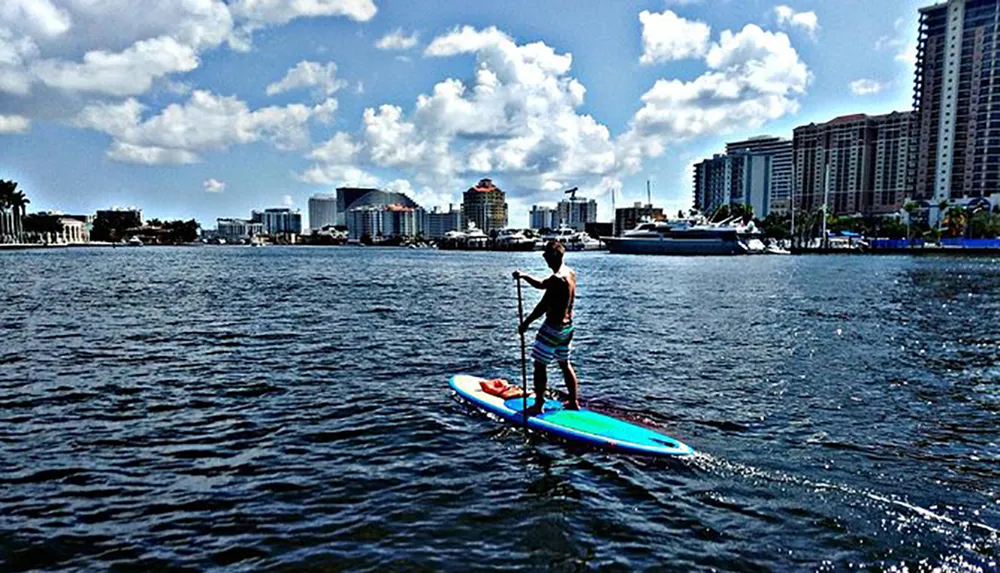 A person is paddleboarding on a calm water body with an urban skyline in the background