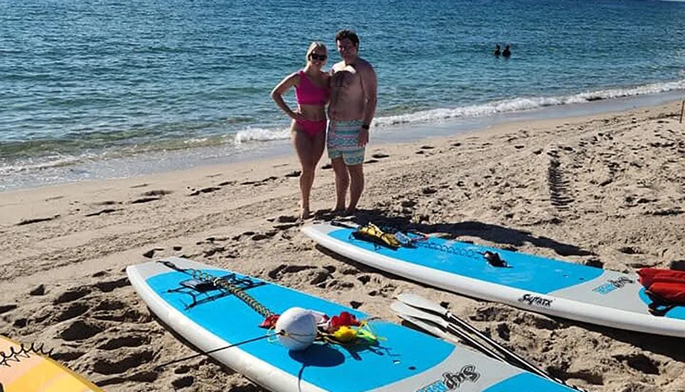 Two people are smiling for a photo on a sunny beach with paddleboards at their feet
