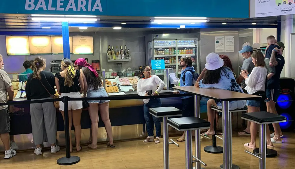 Customers are waiting in line at a Balearia snack bar where donuts and drinks are sold with metal stools and high tables available for seating