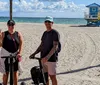 A man and a woman are smiling for the camera while standing next to a Segway on a sunny beach promenade with palm trees and a lifeguard hut in the background