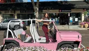 A person is posing with a smile on a parked, pink, open-topped vehicle on a sunny day outside of a shopping area.