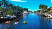 The image shows a tranquil residential canal lined with palm trees and boats docked at private piers, under a clear blue sky.