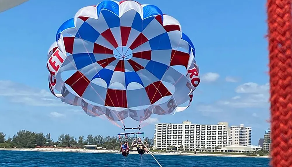 Two people are parasailing over the ocean near a beachfront with buildings in the background under a colorful parachute