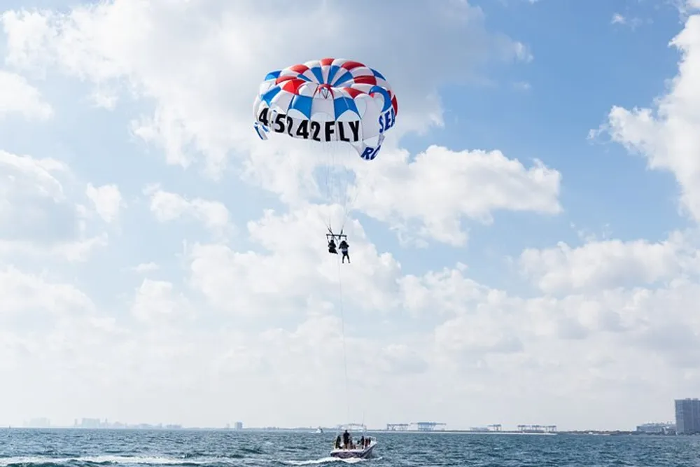 A parasail with two people aloft is tethered to a boat on a body of water under a partly cloudy sky