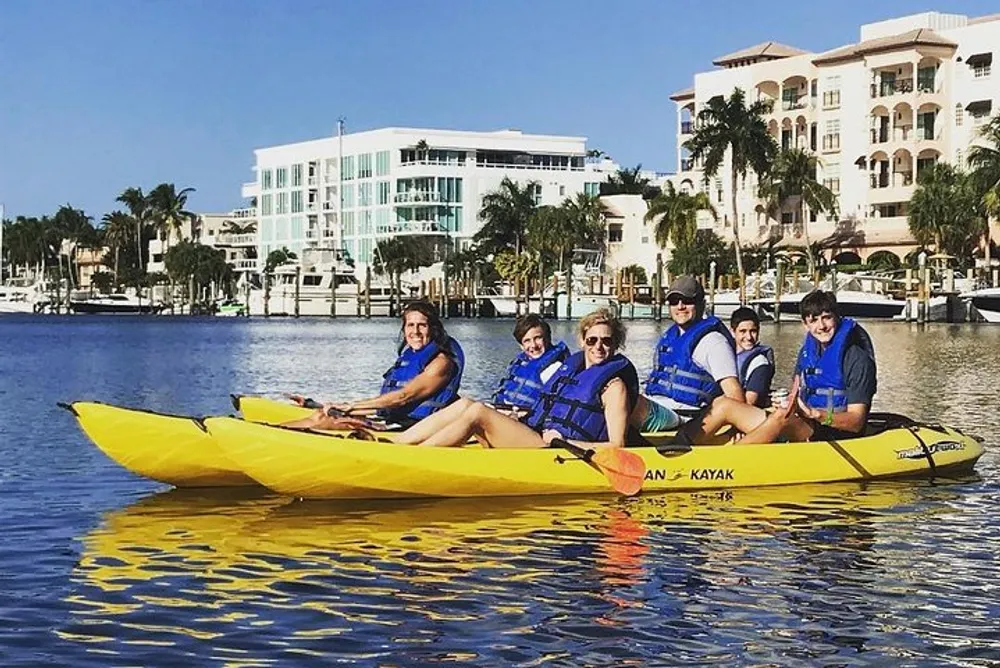 A group of people are smiling and posing for a picture while seated in a tandem kayak on a calm body of water with waterfront buildings in the background