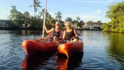 Two people are enjoying kayaking together on a calm waterway lined with palm trees and buildings under a clear sky.