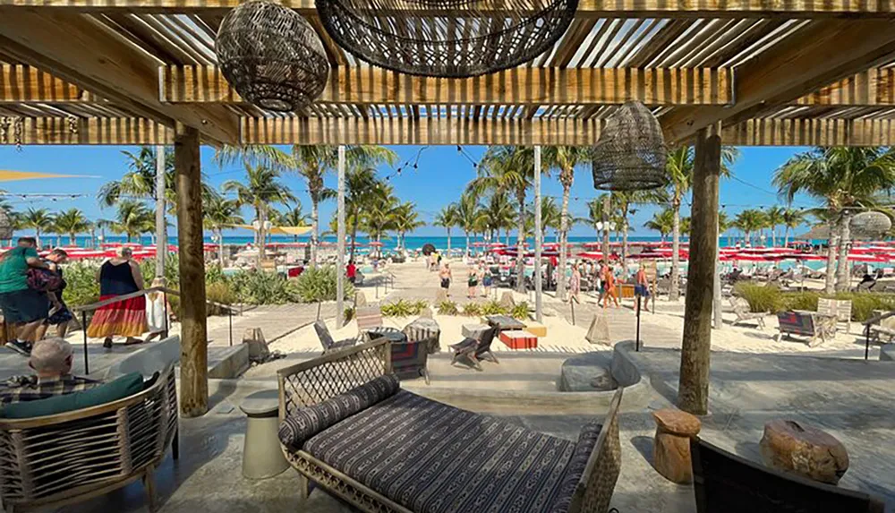 The image shows a tropical beachfront view from a shaded seating area with people enjoying the sunny day amidst palm trees and lounge chairs