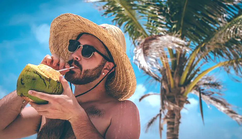 A person is enjoying a drink from a coconut under a sunny sky with palm trees in the background