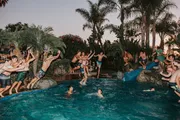 A group of people are enjoying a lively pool party at dusk with several individuals jumping into the water.