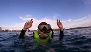 A person wearing a snorkeling mask and a life vest is flashing the peace sign with both hands while floating in the ocean.