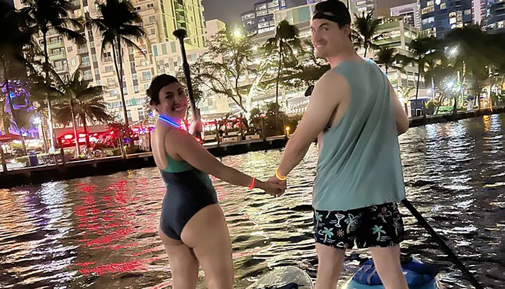 Two people are holding hands and standing on a paddleboard at night with an illuminated cityscape in the background