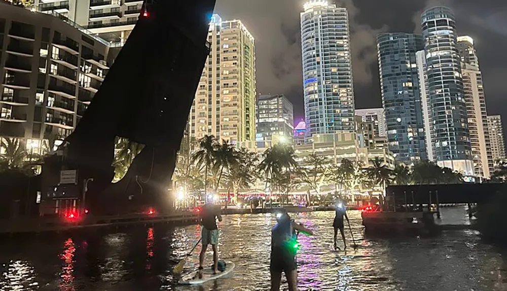 People are paddleboarding at night with illuminated paddleboards against a backdrop of a brightly lit cityscape and raised drawbridge