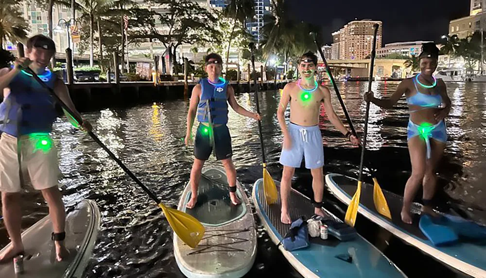 Four people adorned with neon lights stand on paddleboards at night in an urban waterway ready for a glowing aquatic adventure