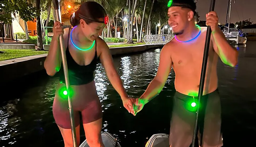 A man and a woman illuminated by glowing necklaces and lights on their paddles and paddleboards hold hands while standing on their boards in a body of water at dusk or night
