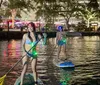 Two individuals are stand-up paddleboarding at night with LED lights on their boards creating a vibrant scene on the water against an urban backdrop