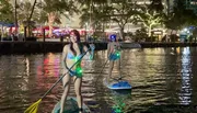 Two individuals are stand-up paddleboarding at night with LED lights on their boards, creating a vibrant scene on the water against an urban backdrop.