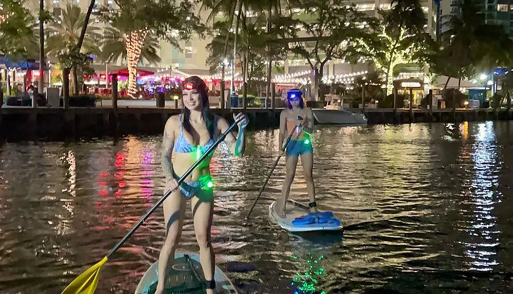 Two individuals are stand-up paddleboarding at night with LED lights on their boards creating a vibrant scene on the water against an urban backdrop
