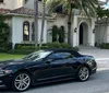 A black convertible car is parked outside a luxurious two-story house with palm trees and a clay tile roof