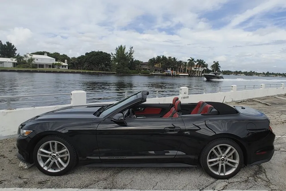 A black convertible car with red interior is parked by a waterfront with tropical vegetation in the background