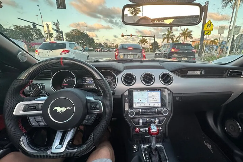 A drivers view from inside a Mustang displaying the dashboard steering wheel and center console with a view of other cars on the road ahead at twilight