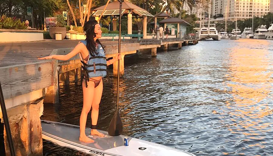 A person is standing on a paddleboard near a dock during sunset, with the golden light reflecting off the water.
