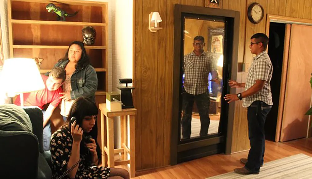 A group of people is seen inside a warmly lit room with one person talking on the phone another standing by the door with their reflection visible and two individuals appearing to be in a conversation behind a lamp